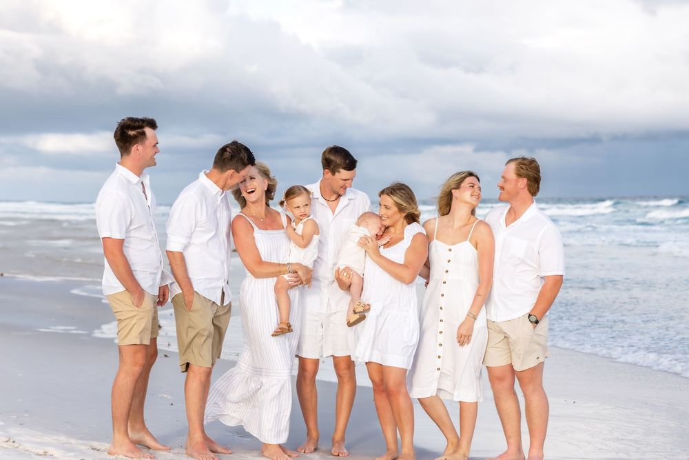 A group in coordinated white outfits and khaki shorts poses together on a sandy beach by the ocean.