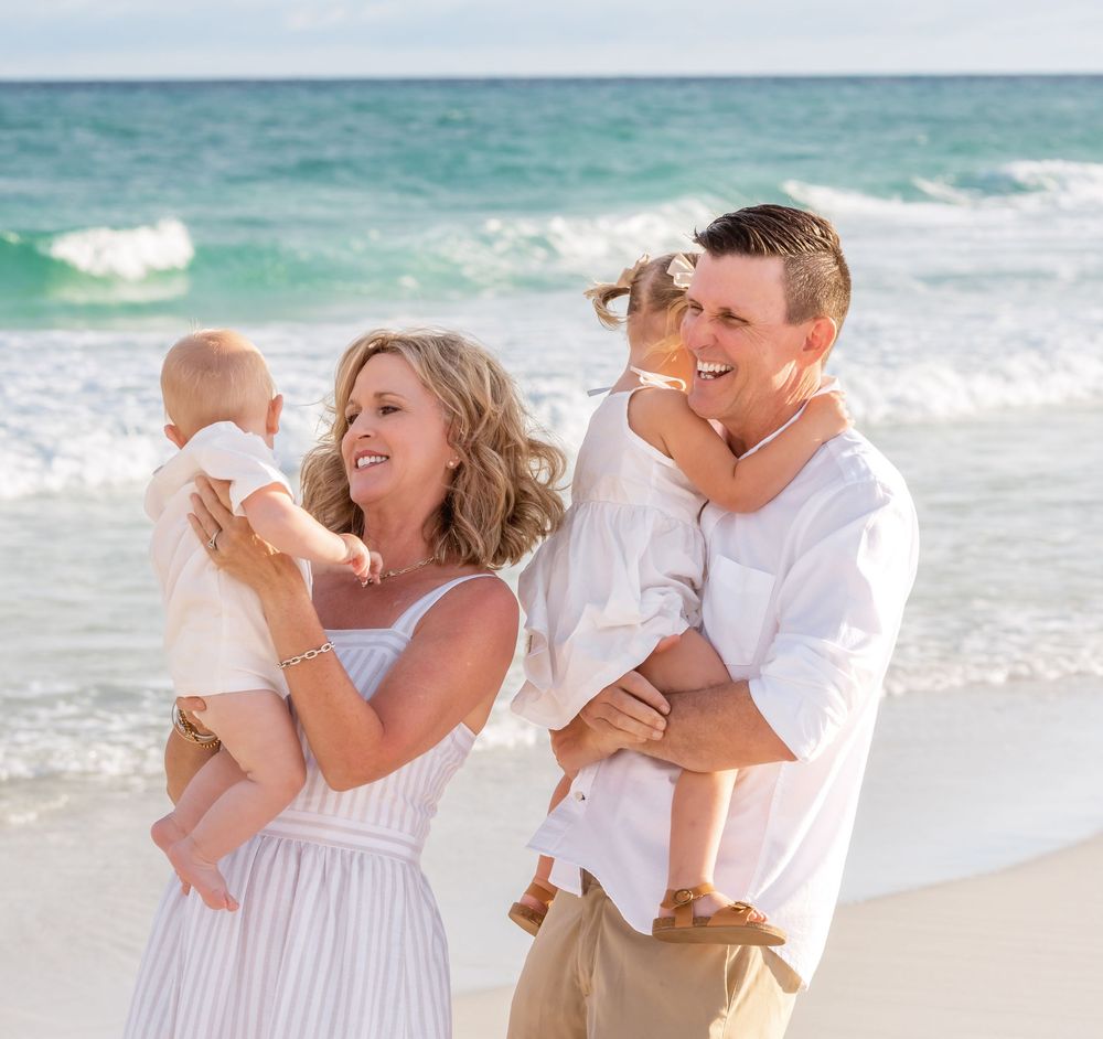 A happy family in white outfits enjoys a tender moment together while standing on a sunny beach with ocean waves.