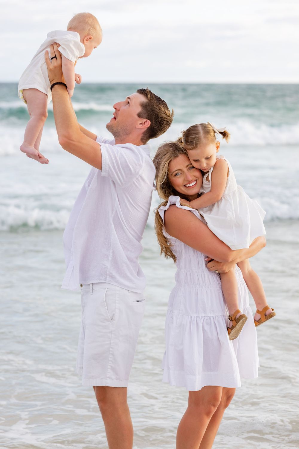Family wearing all white outfits playing together on a sunny beach with ocean waves in the background.