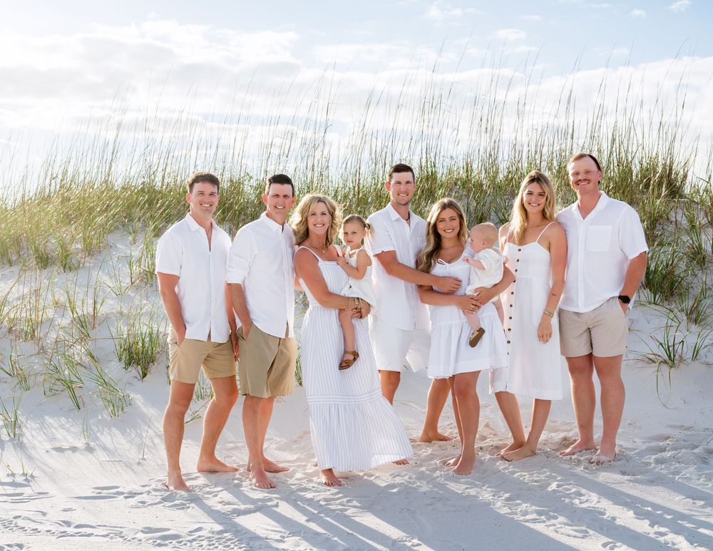 A group wearing white and beige clothing poses together on a sunny beach with sand dunes and seagrass in background.