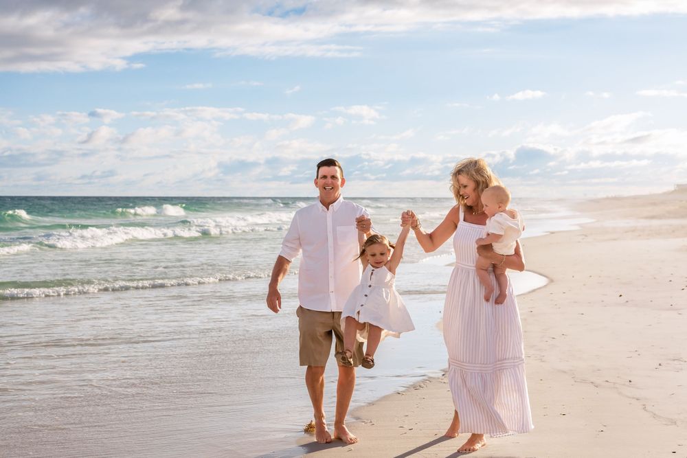 A family walks along pristine white sand beach with ocean waves and cloudy sky in the background.