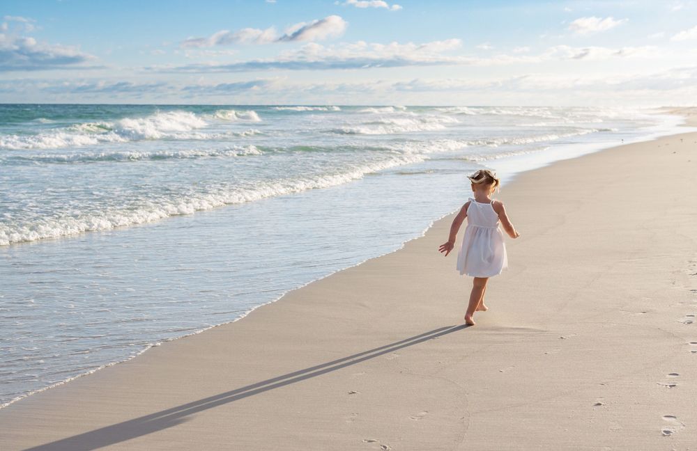 Small child in white dress runs joyfully along empty beach shoreline during golden hour.