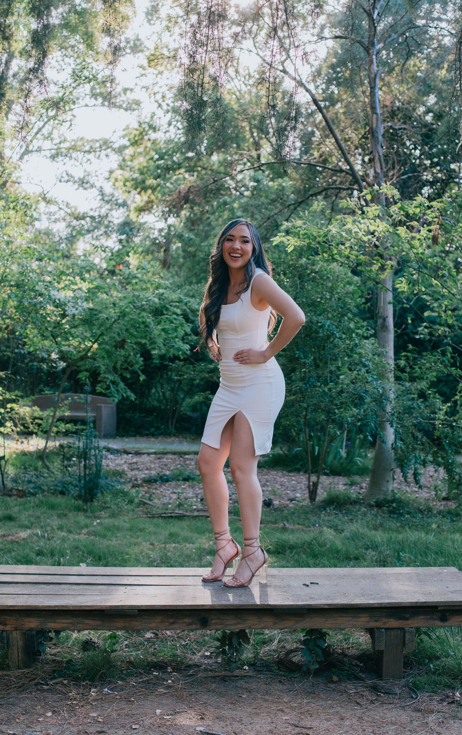 Someone in a white dress strikes a pose on a wooden deck platform in a lush green forest setting.