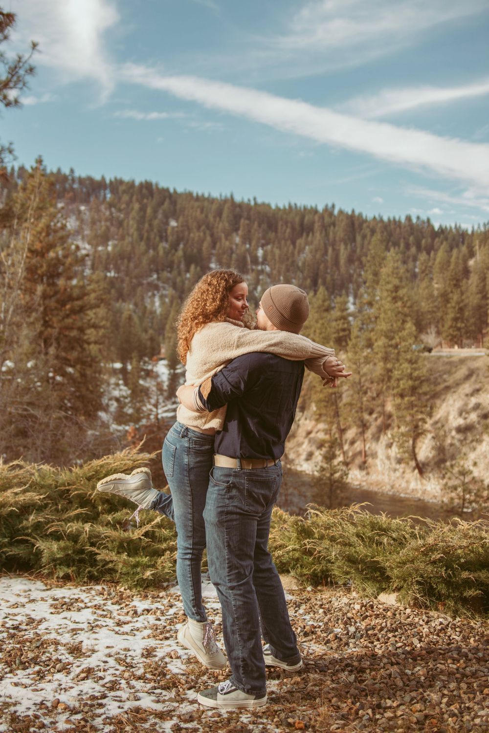 Couple embracing on a snowy mountain trail with evergreen forest and blue sky in the background.