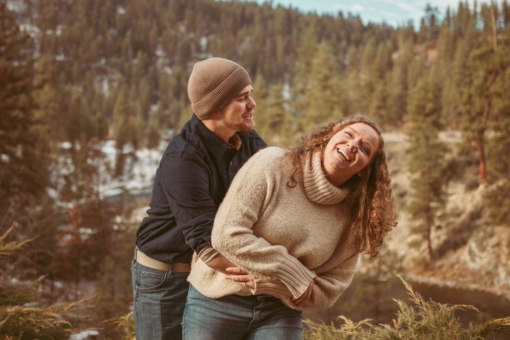 Two people enjoying a playful moment outdoors in a forest setting with mountains in the background on a sunny day.