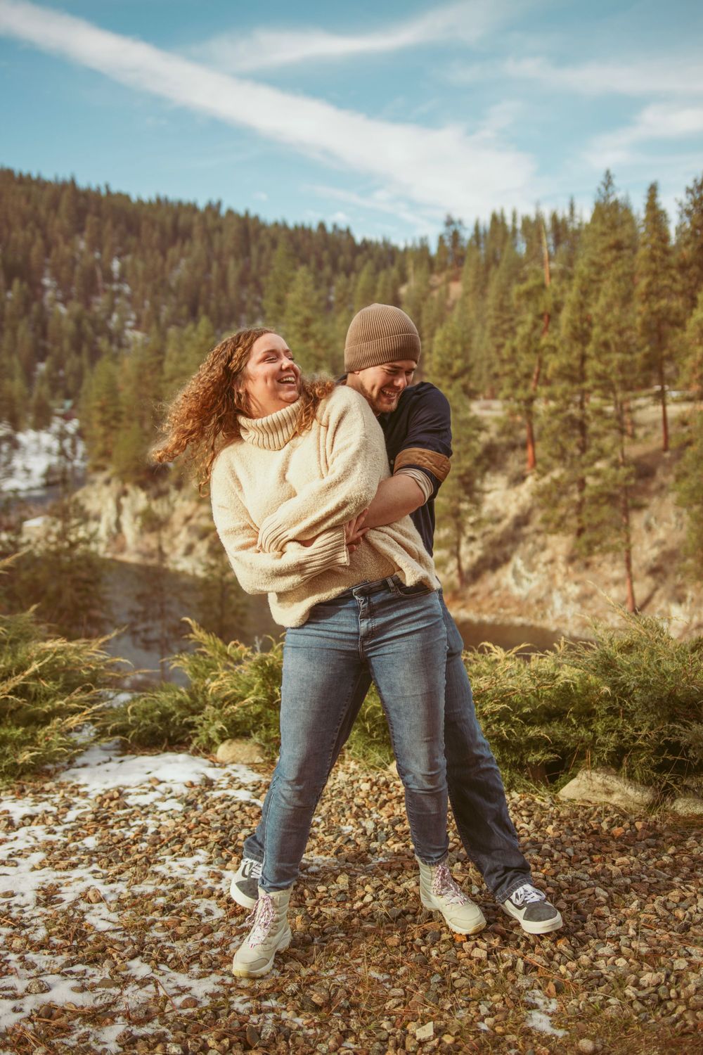 A couple shares a joyful moment while embracing on a mountainside with evergreen forest and snowy slopes in background.
