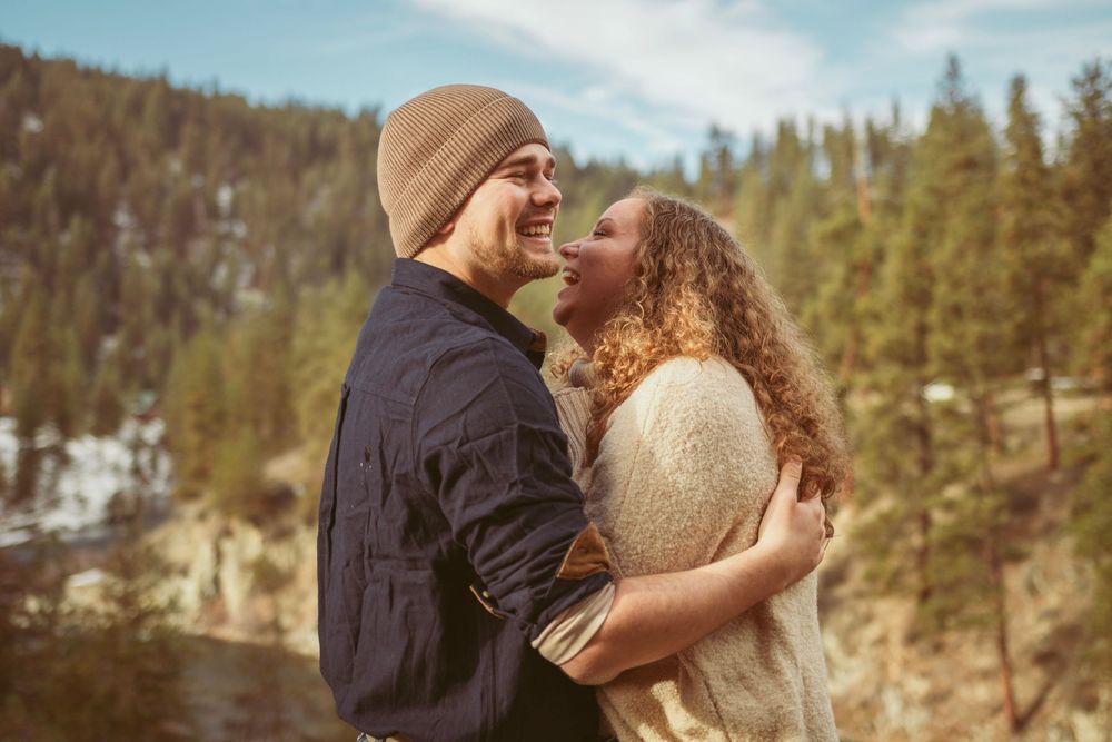 A joyful couple embrace and laugh together against a scenic mountain forest backdrop on a winter day.