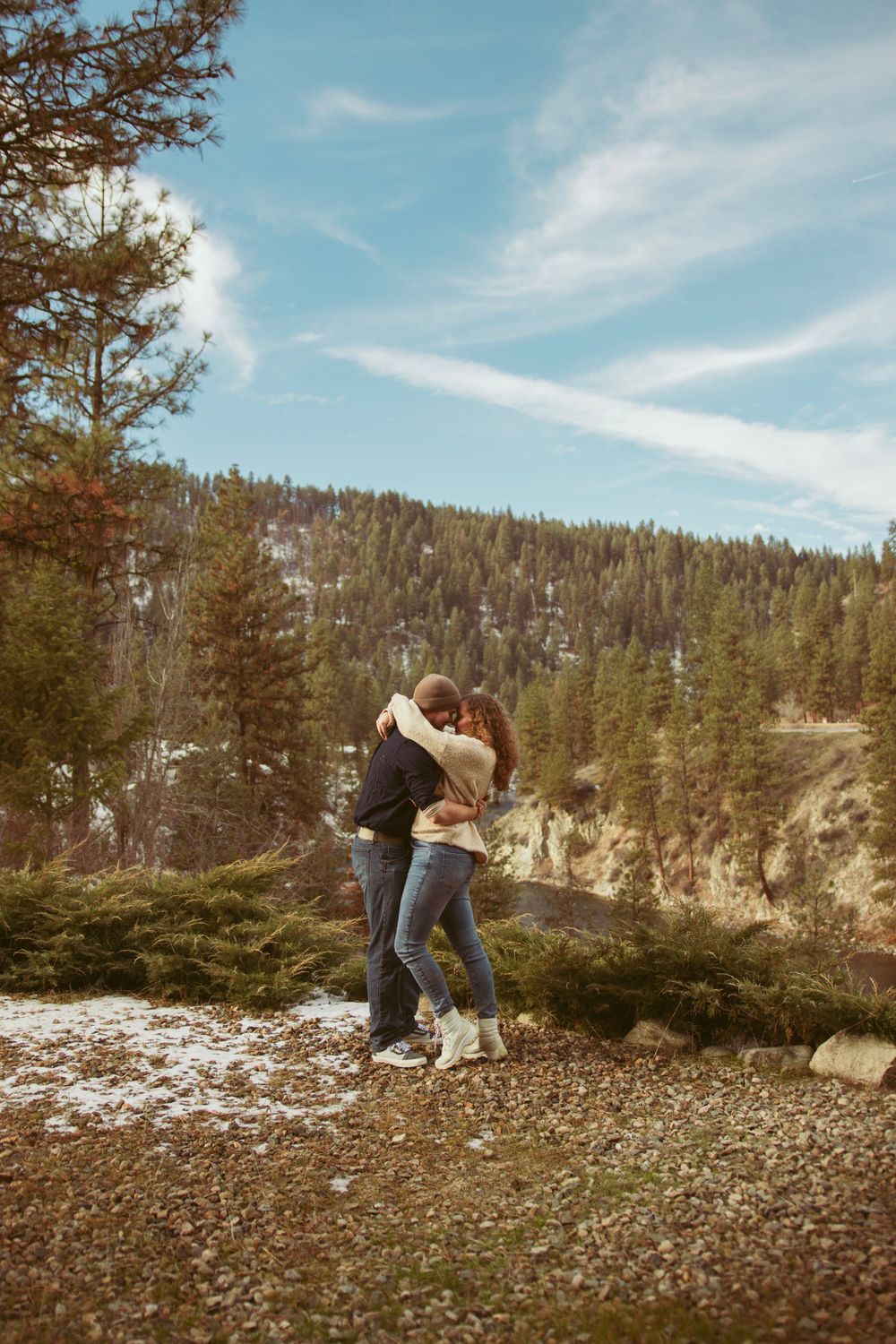 Two people embrace in a romantic moment on a snowy mountainside with evergreen forest and cloudy blue skies in the background.