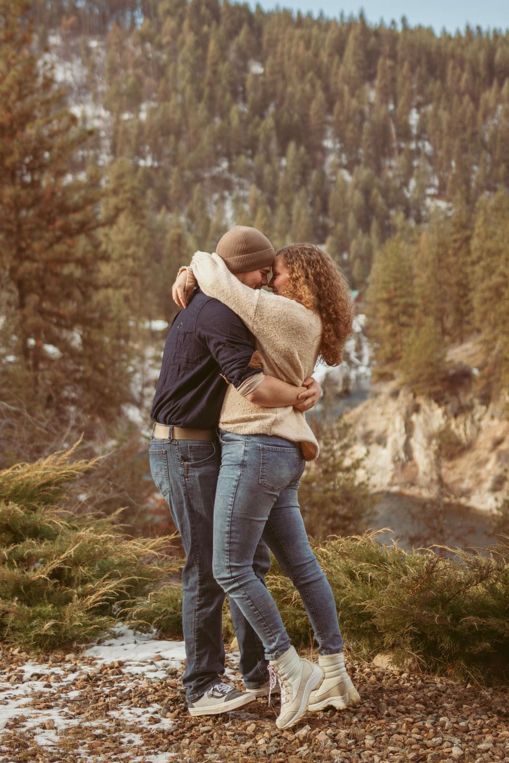Two people playfully embrace on a snowy hillside with evergreen forest and mountain scenery in the background.