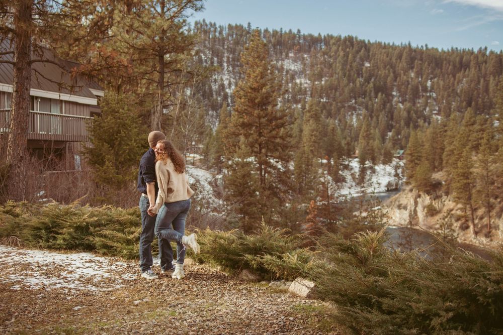 A couple stands on a snowy mountainside trail with pine trees and a mountainous landscape in the background.