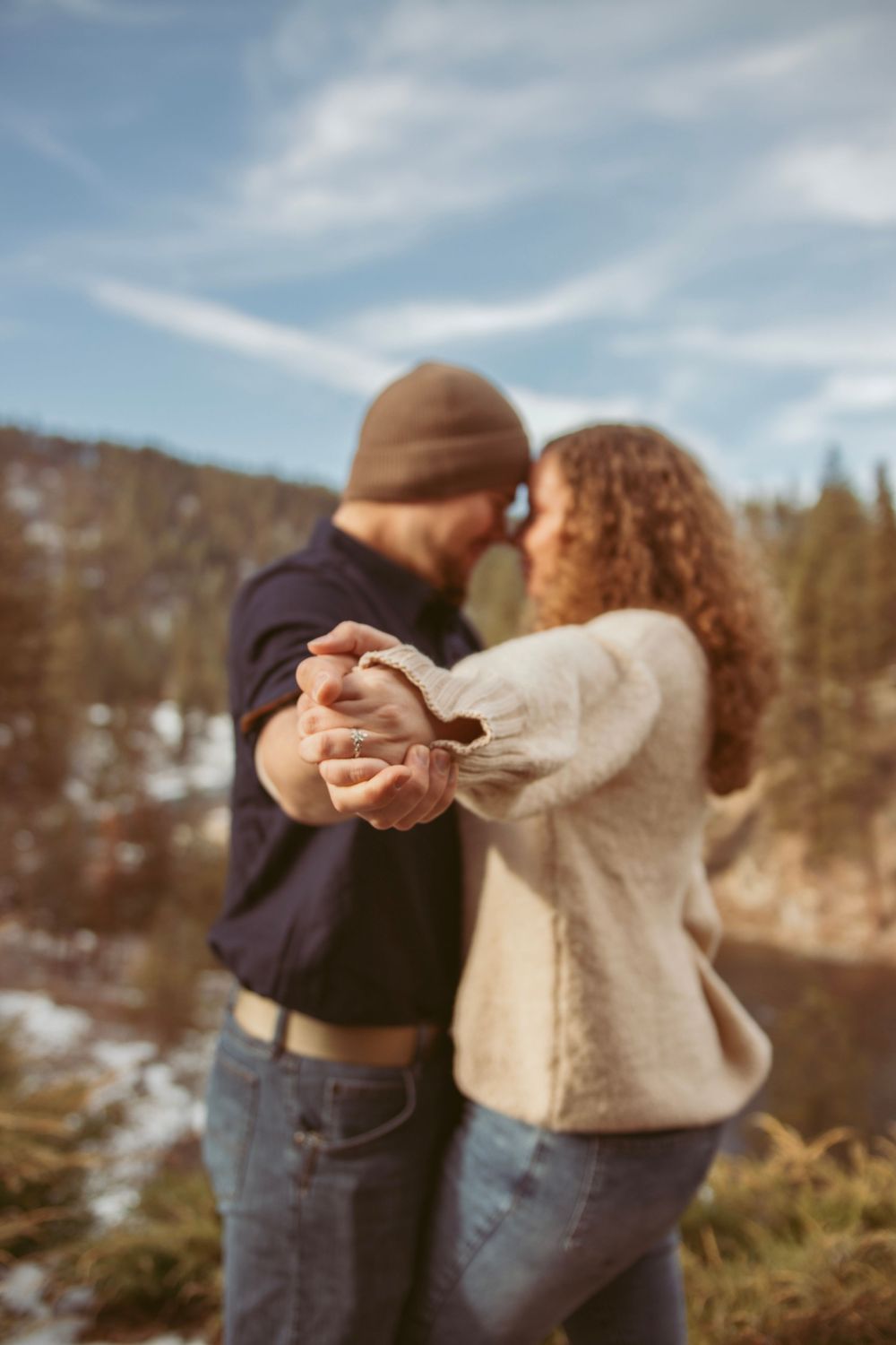 A couple dances together on a mountain overlook with pine trees and snow-covered slopes in the distance.