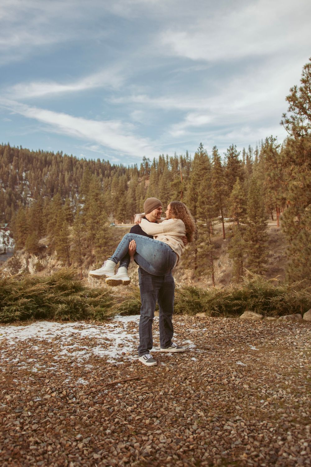 A couple embraces against a picturesque mountain landscape with pine trees and light snow on the ground.