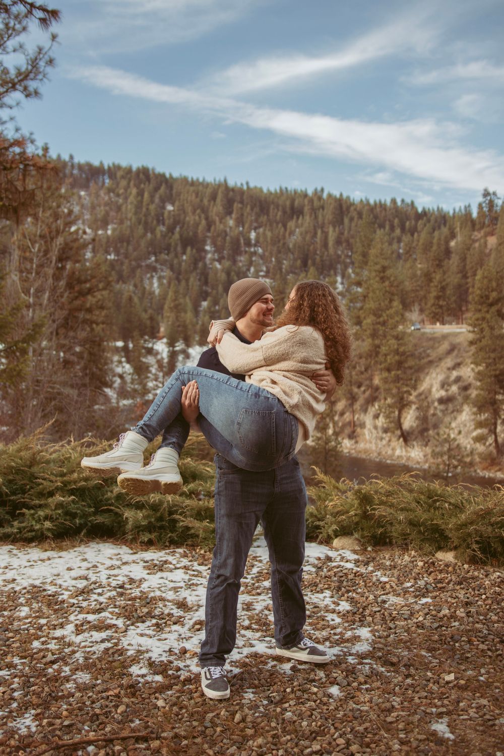 A couple in winter attire dances together in front of a snowy mountain landscape during a photoshoot.
