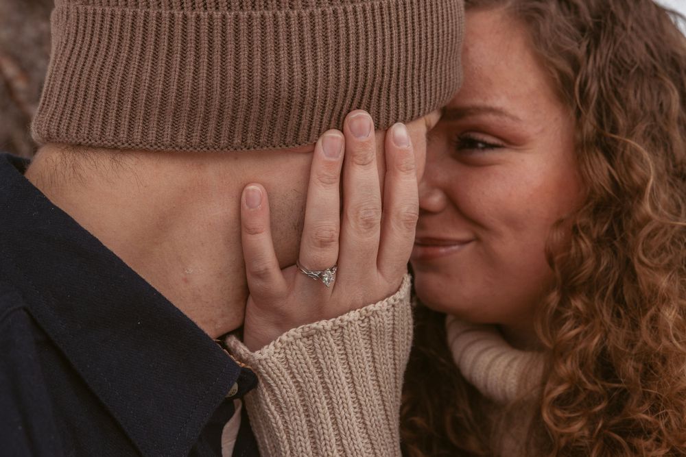 Someone with curly hair covers their mouth while sharing an intimate moment with their partner in cozy sweaters.