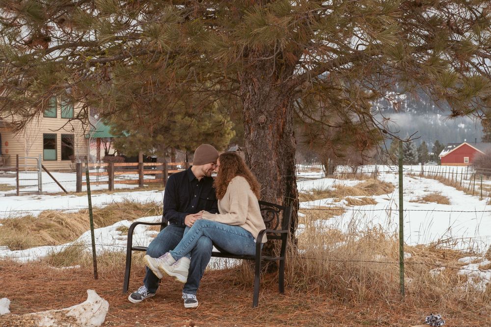 A couple sits intimately on a wooden bench under a snow-dusted tree with rustic farm buildings in the background.