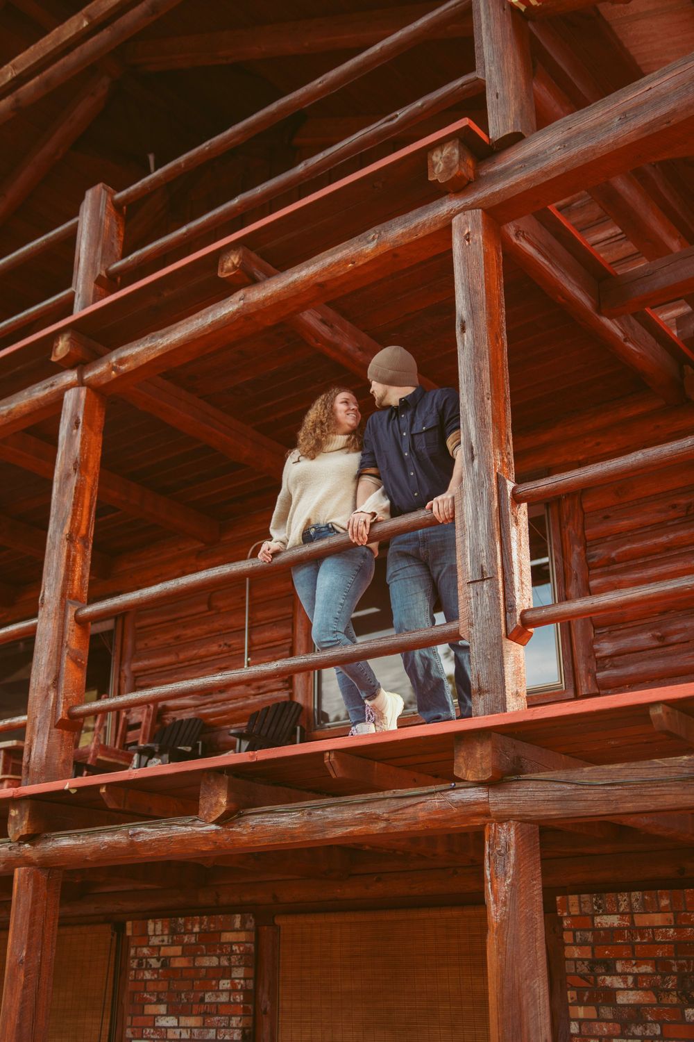 Two people lean against wooden railing on rustic cabin porch with exposed timber beams and brick walls.