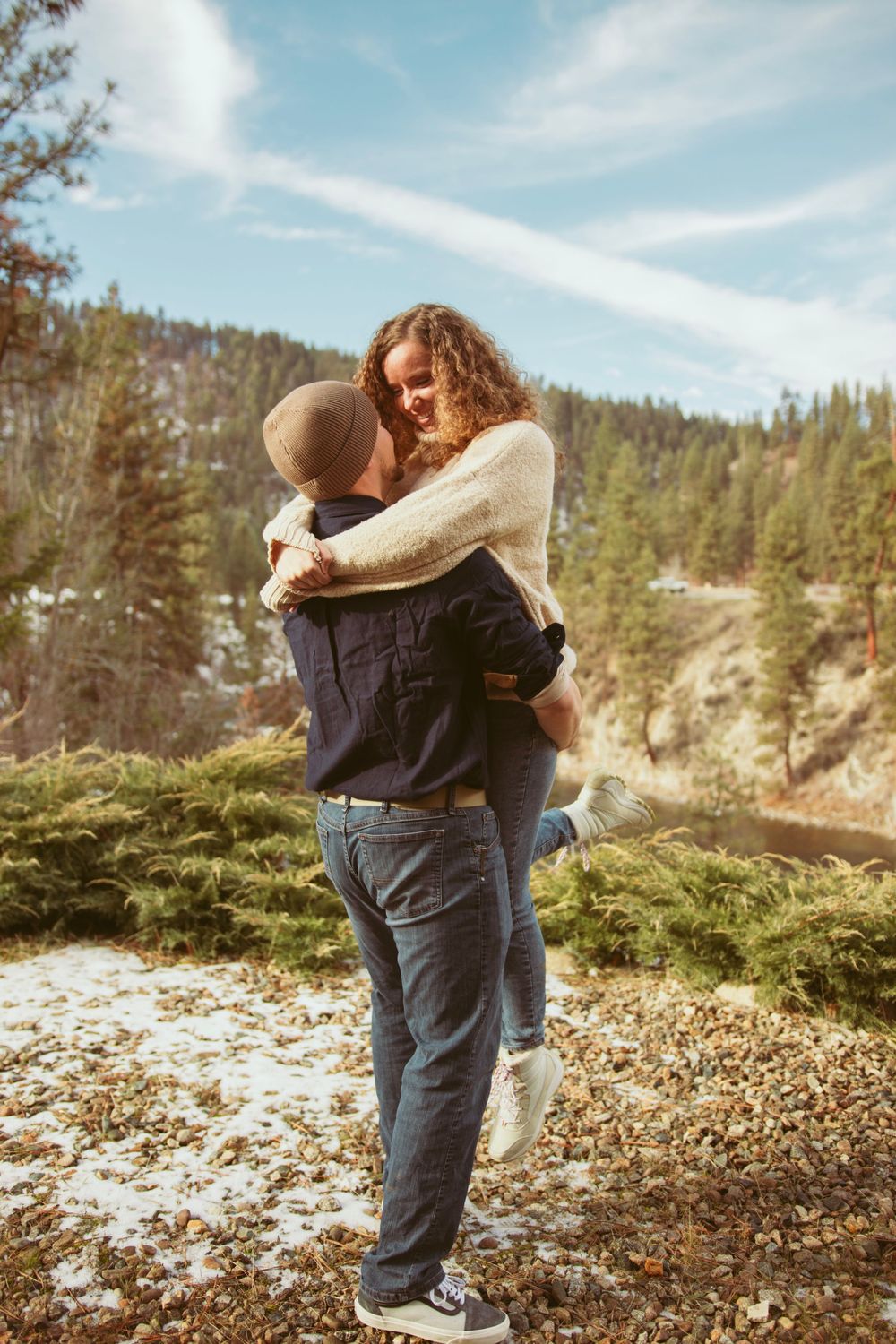 Romantic couple embraces against scenic mountain backdrop with pine trees and blue skies.