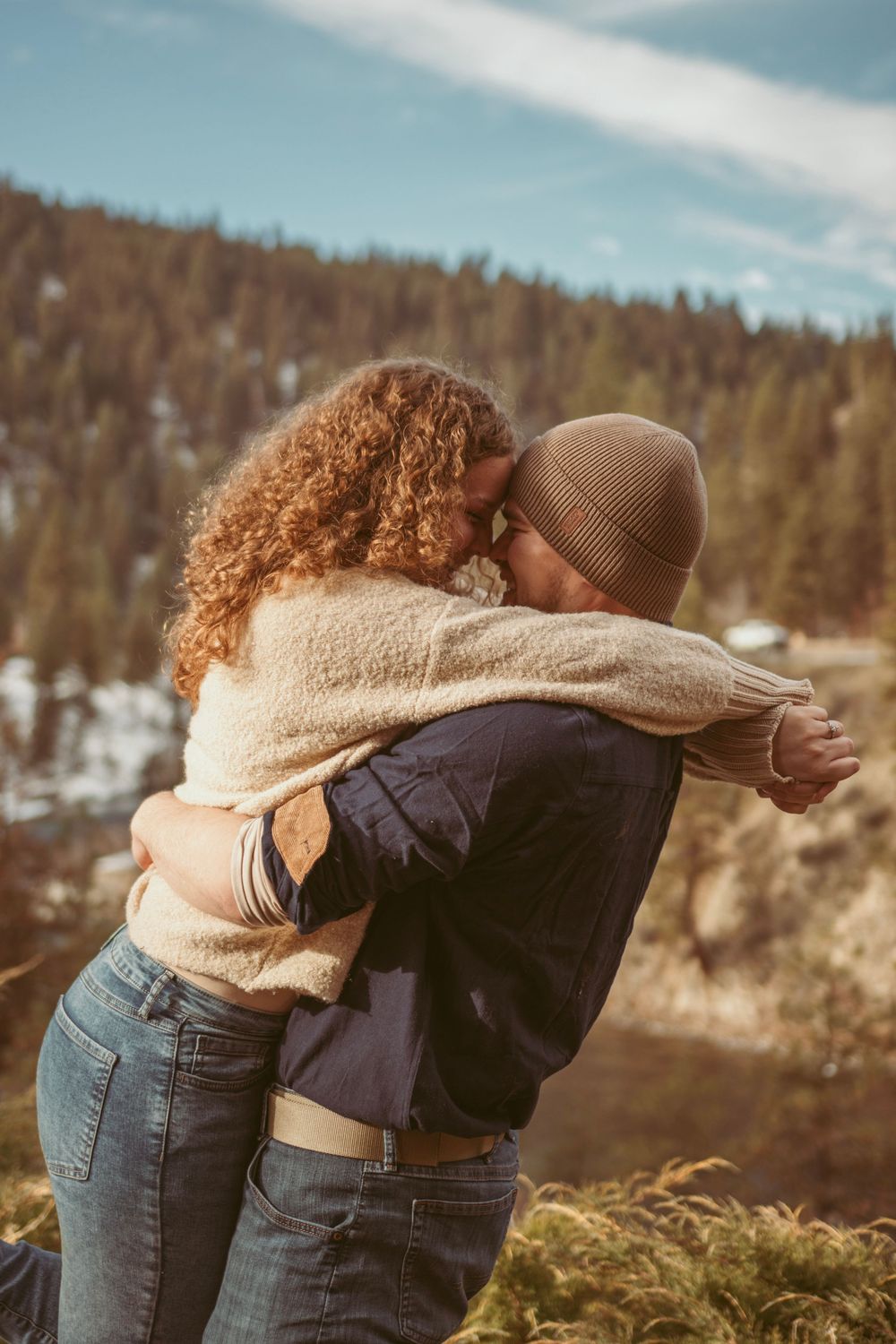 Two people embracing on a mountainside with snow covered hills and forest in the background on a sunny day.
