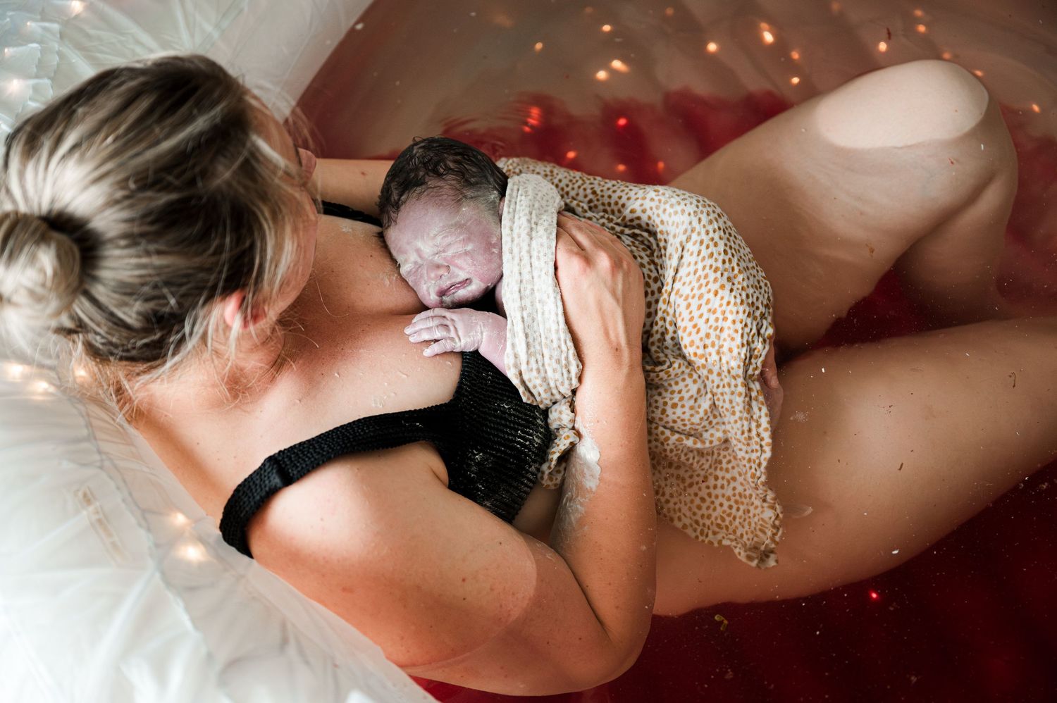 A tender moment with a newborn baby swaddled in a knit blanket against someone lying on white bedding.
