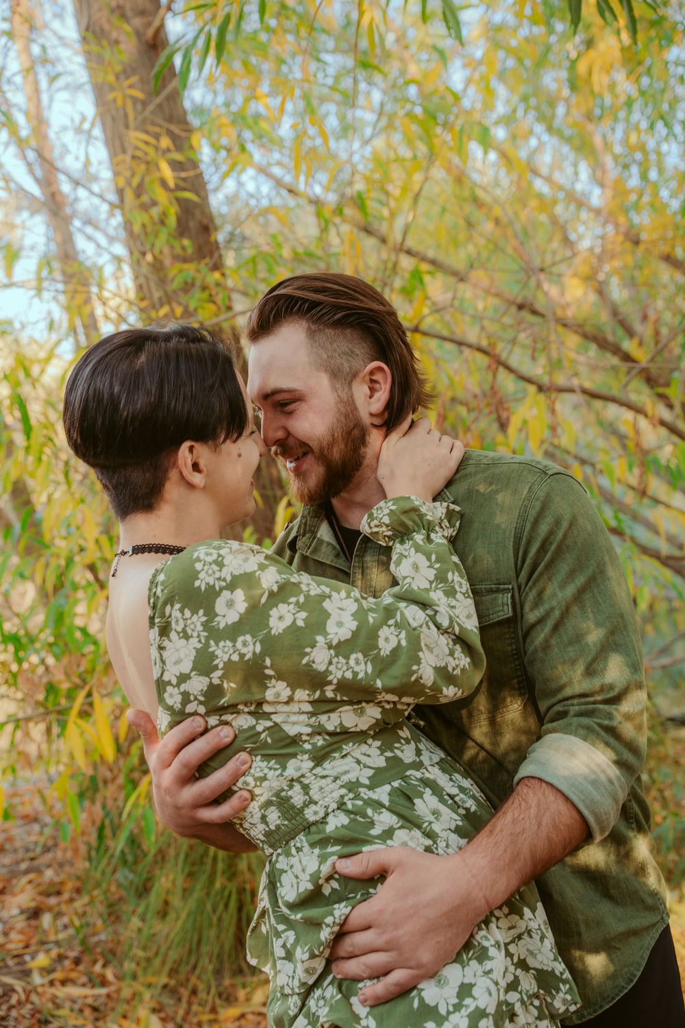 Couple sharing an intimate embrace outdoors on an autumn day with golden willow trees in background.