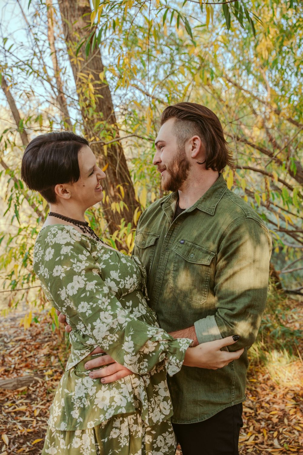 Couple embracing outdoors under a willow tree with golden sunlight filtering through the branches in autumn.