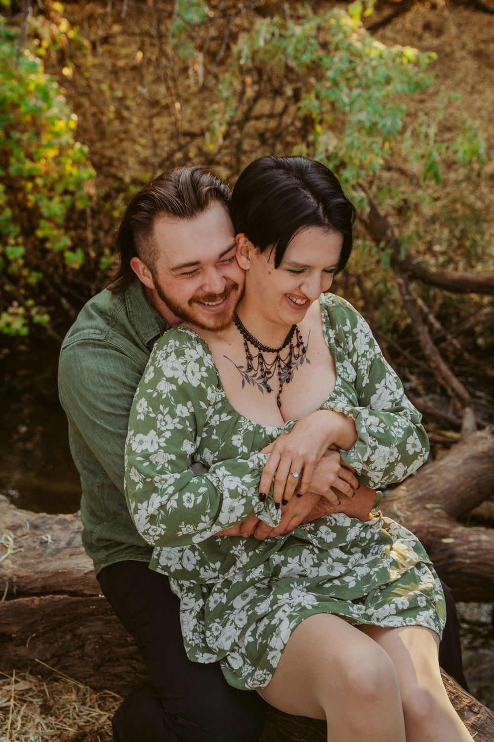 A couple shares an intimate moment outdoors on a fallen log, wearing green clothing amid autumn foliage.