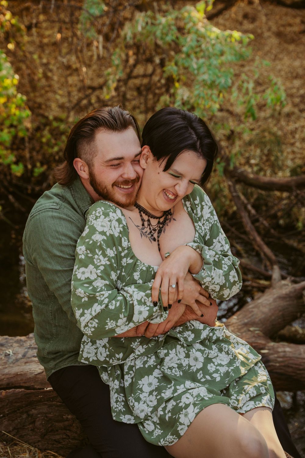 A romantic couple shares a tender embrace while sitting on a fallen log in a wooded setting.