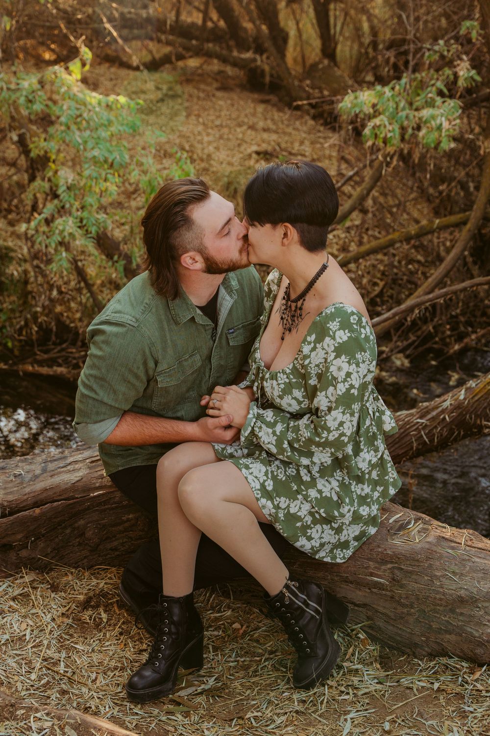 Romantic couple sits on fallen tree trunk in autumn forest setting, sharing intimate moment while wearing casual attire.