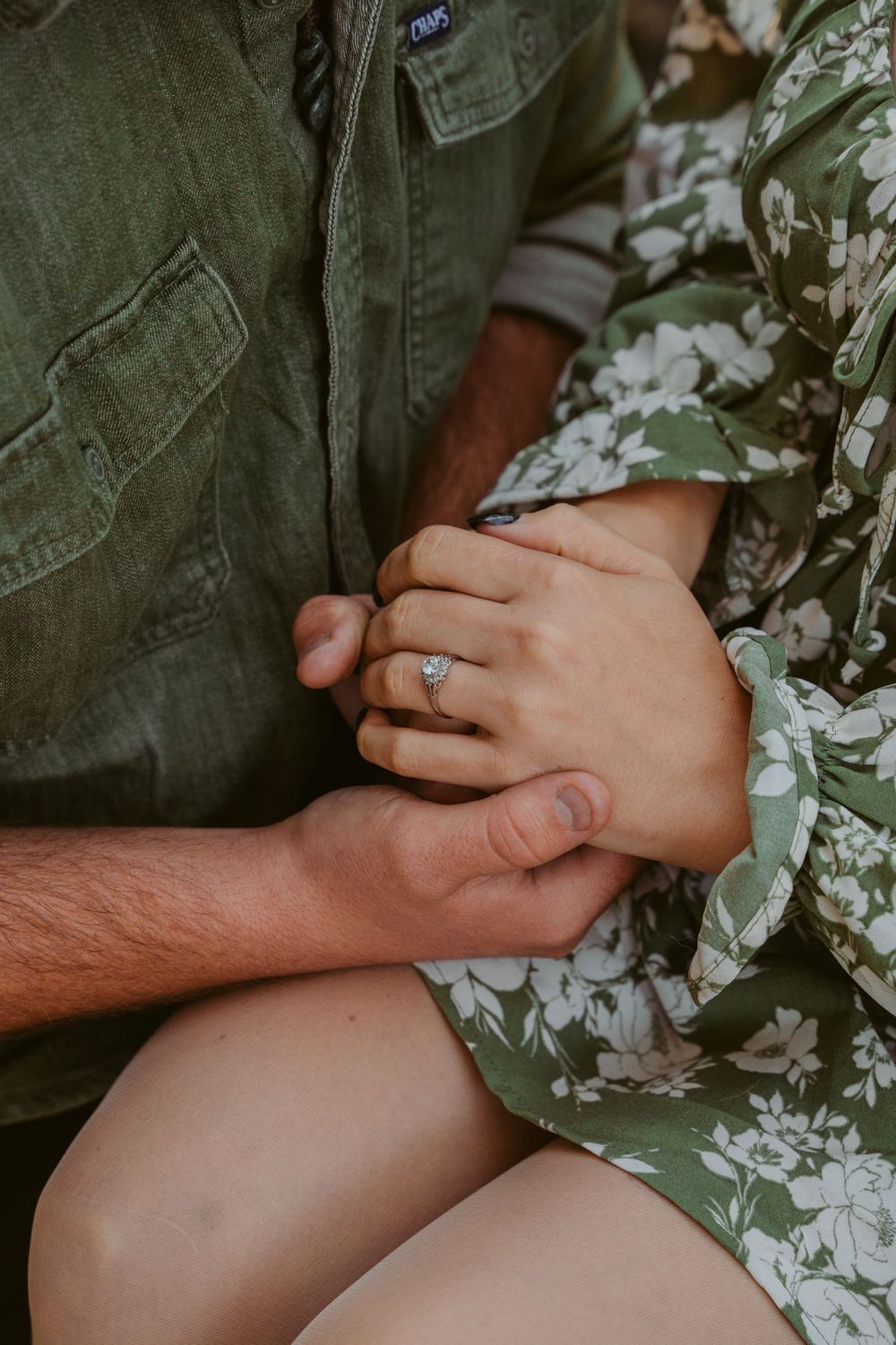 Close up of hands holding, with an engagement ring visible on floral green dress against olive jacket.
