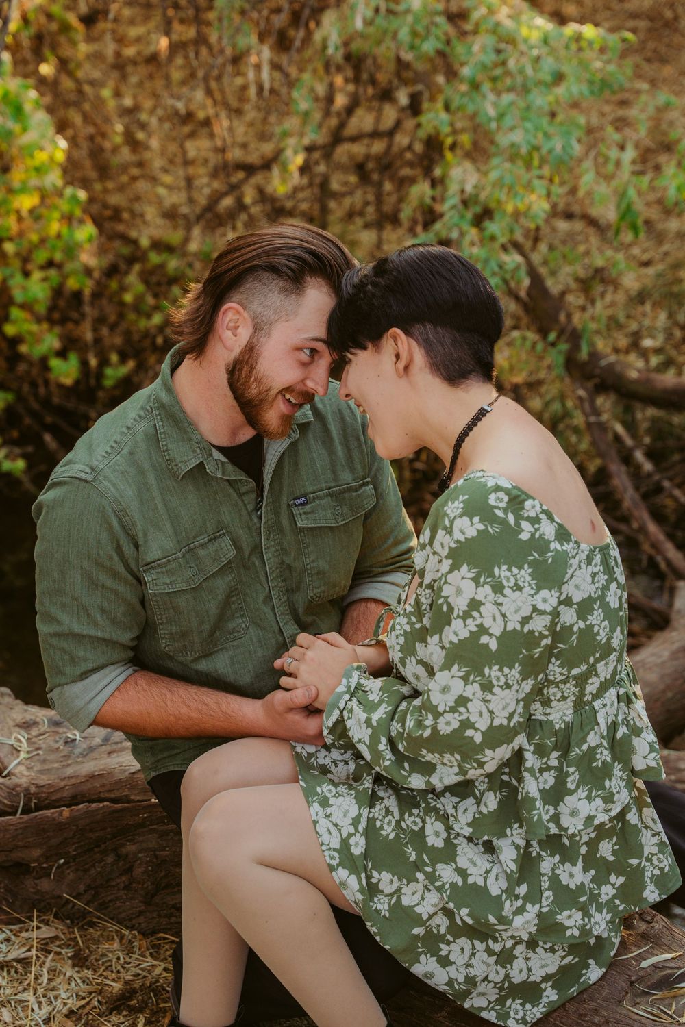 A couple in coordinating green outfits share an intimate moment while sitting together outdoors in nature.