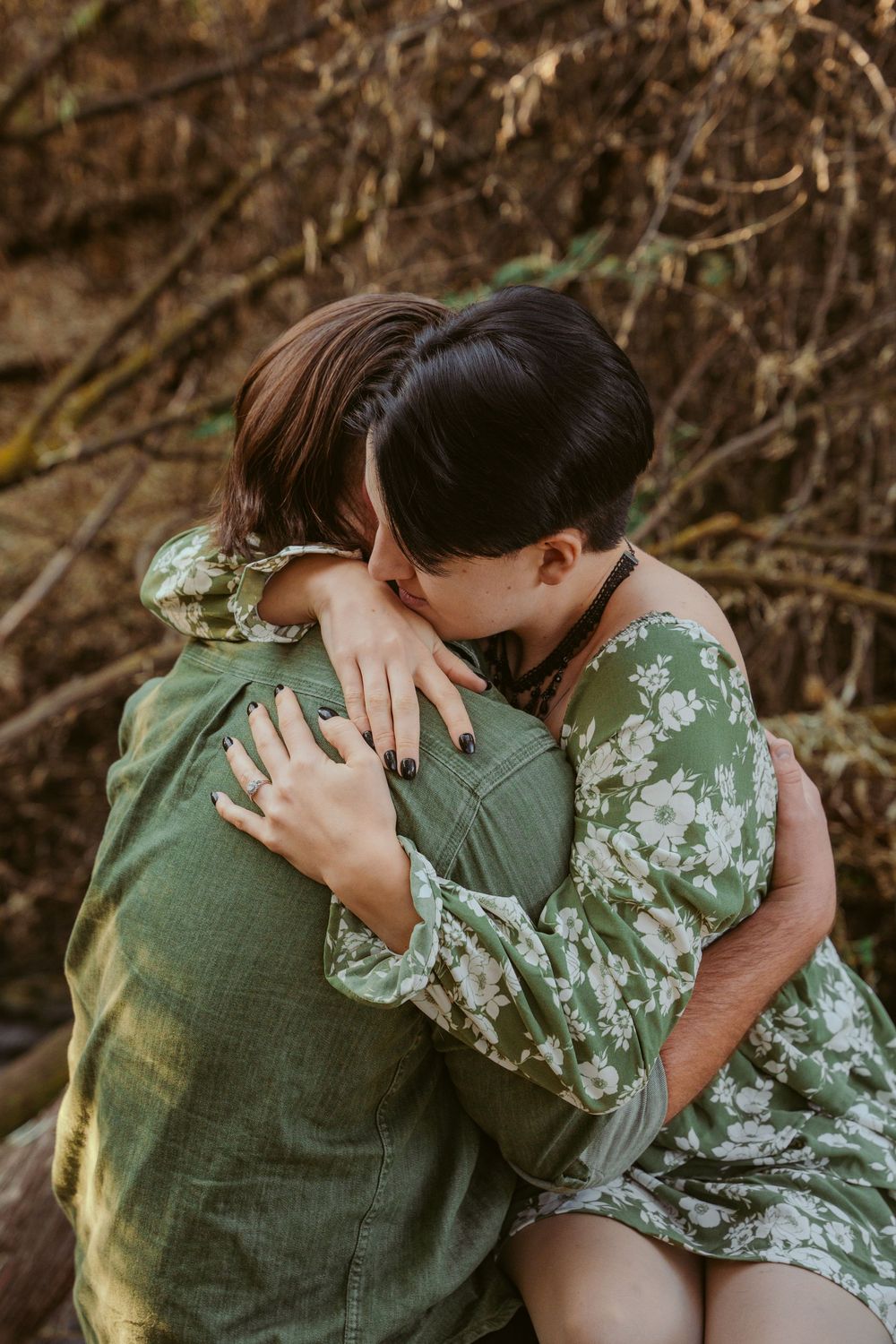 Two people embrace tenderly in a romantic moment, one wearing a green floral dress against a natural outdoor backdrop.
