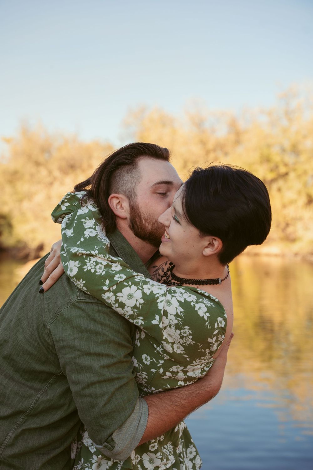 A romantic couple shares tender kisses by the waterfront at sunset while wearing coordinating green outfits.