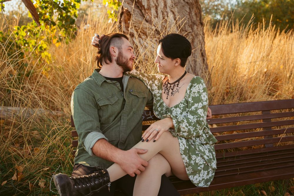 A couple sits close together on a wooden bench in a sunlit field with tall golden grass and trees in the background.
