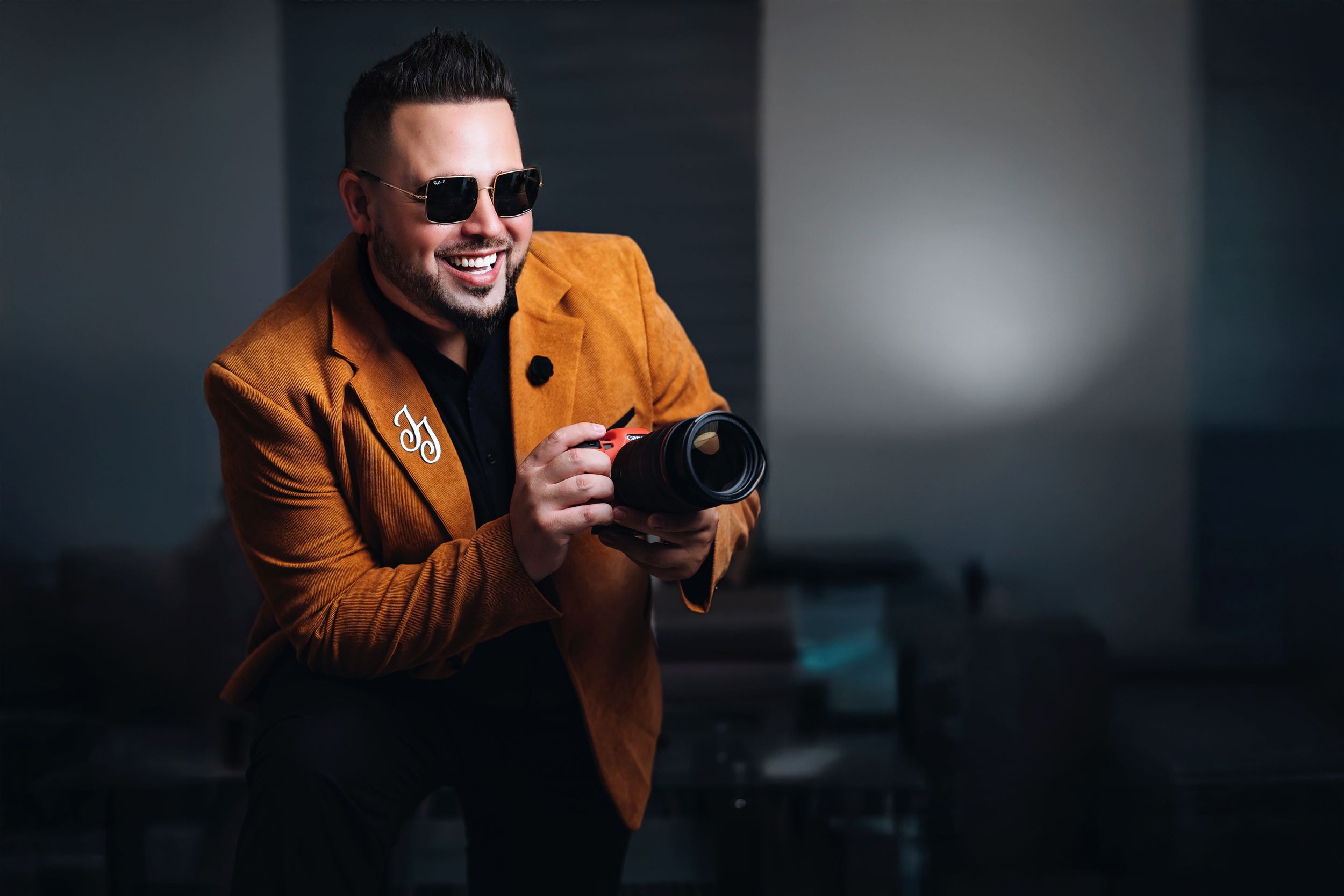 Person in orange blazer and sunglasses looking at smartphone with joyful expression in dark studio setting.