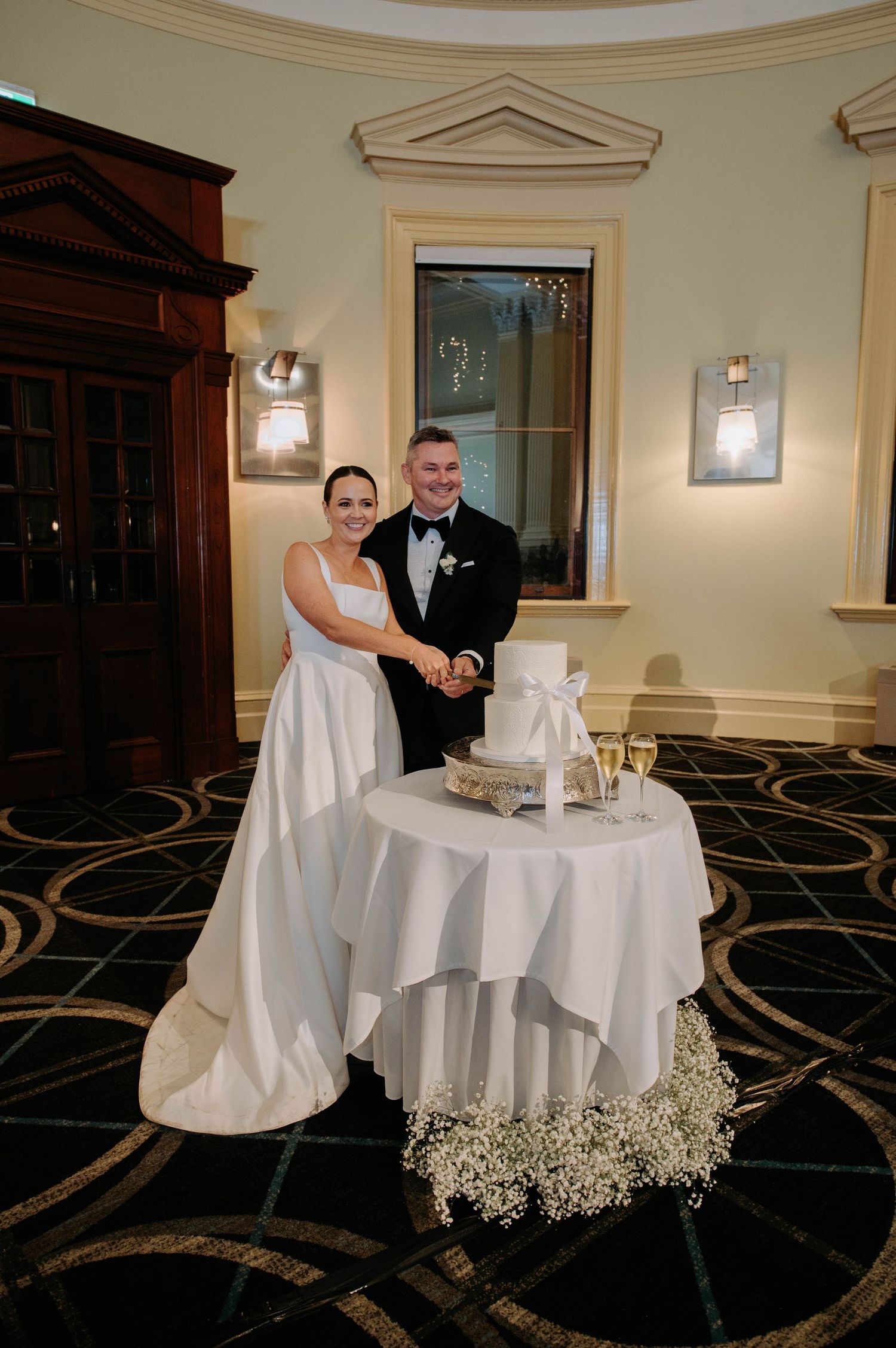 A wedding couple stands beside their elegant white wedding cake on a pedestal table decorated with baby's breath flowers.
