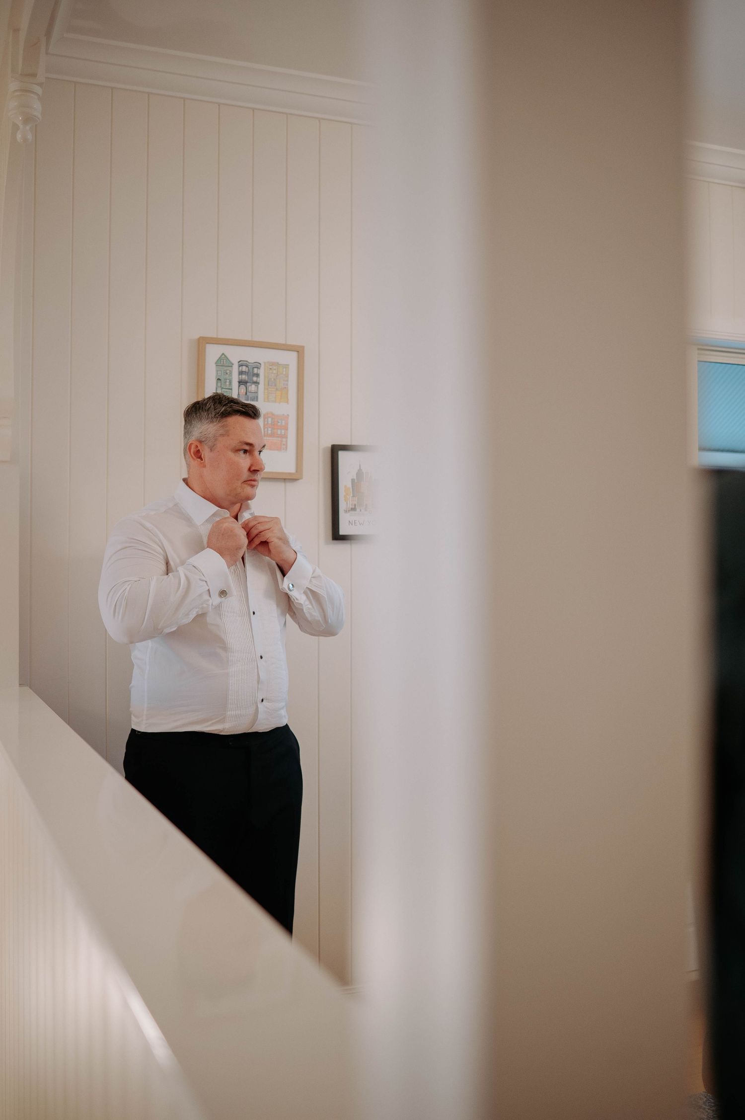 A person in formal attire adjusts their tie while standing near a white staircase in an elegant interior setting.