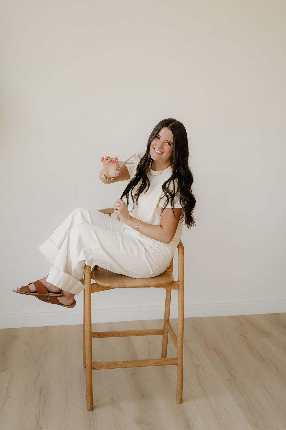 Person in white outfit sitting casually on wooden stool against neutral wall with light hardwood flooring.