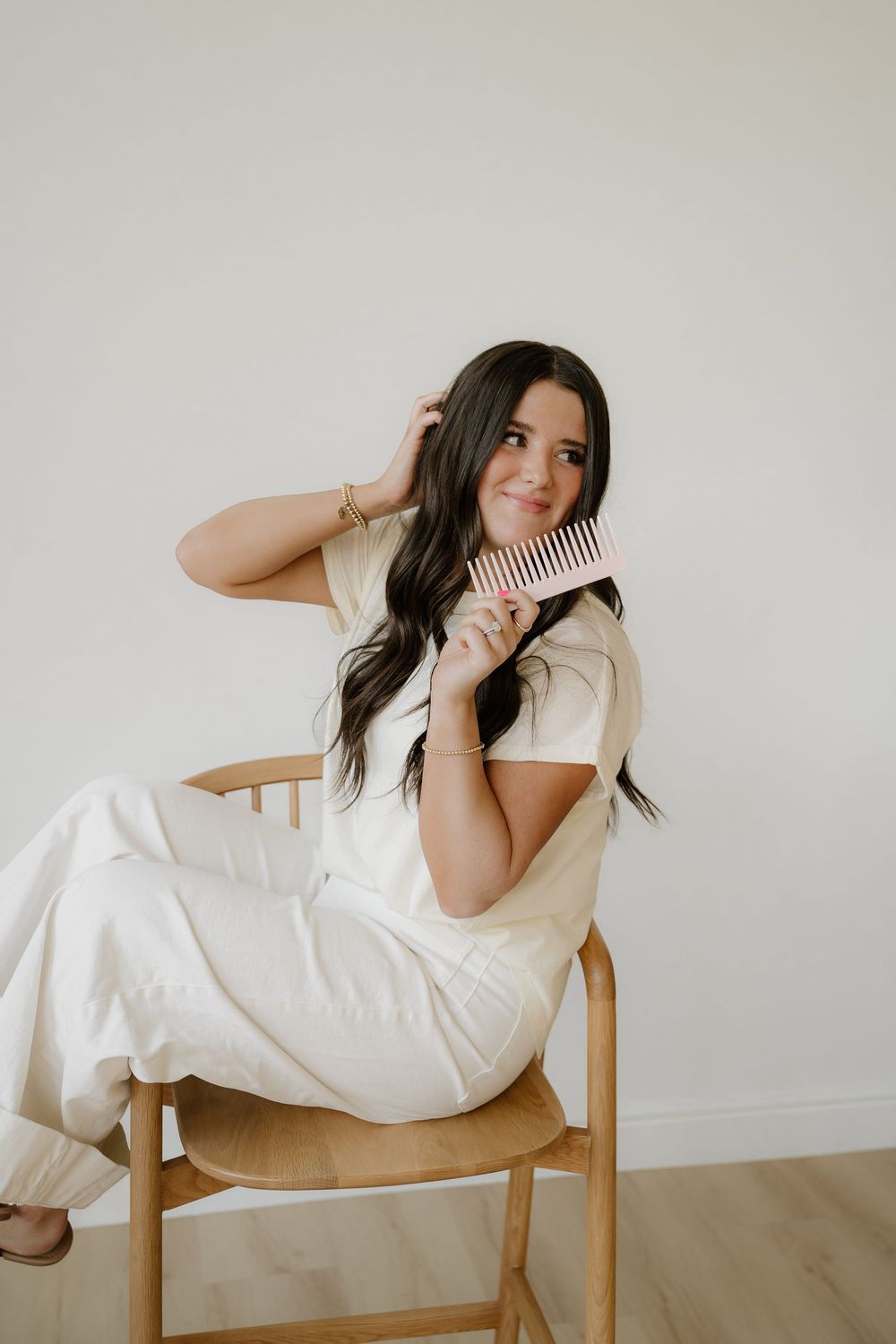 A person in a cream-colored outfit sits casually on a wooden chair holding a hairbrush against a neutral wall.