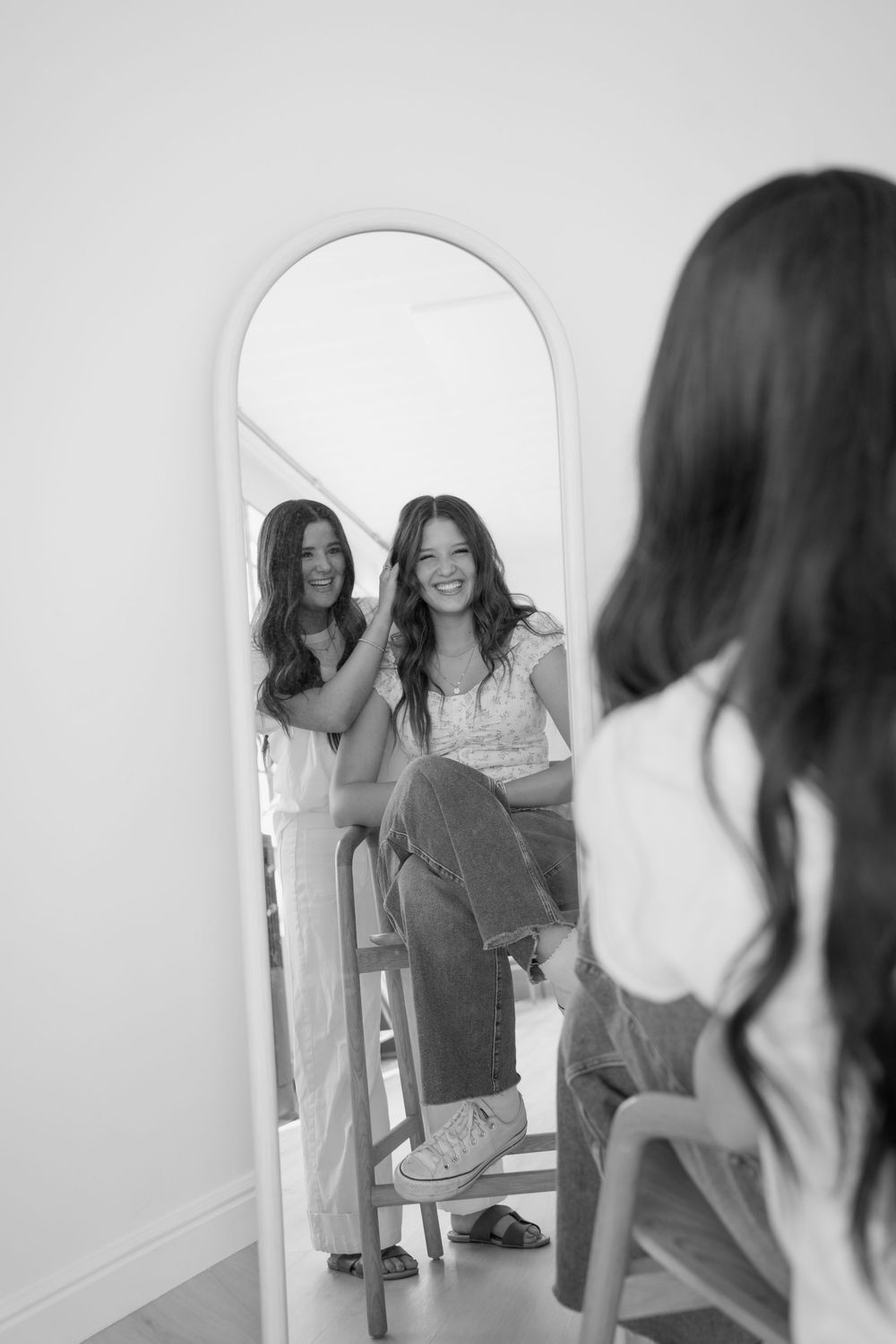 Black and white photo showing reflection in an arched mirror of someone sitting on a chair wearing jeans and white top.
