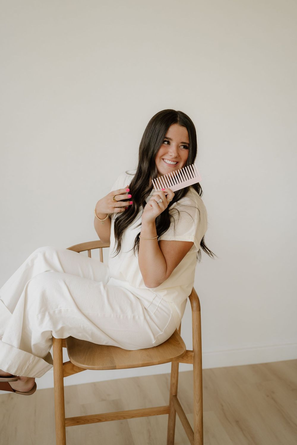 A person in a white outfit sits casually on a wooden chair holding a pink hair comb and smiling.
