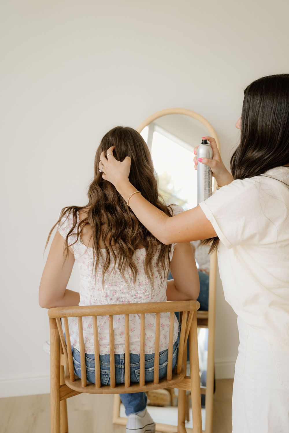 Hair styling session in progress with person seated in wooden chair near arched mirror.