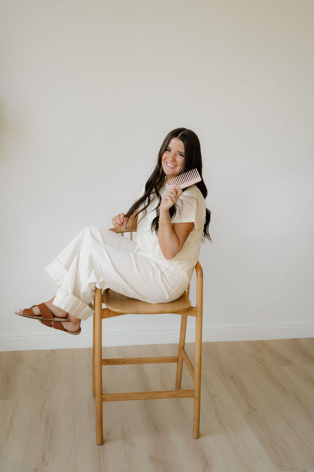 Woman in white outfit posed casually on wooden stool against neutral wall.