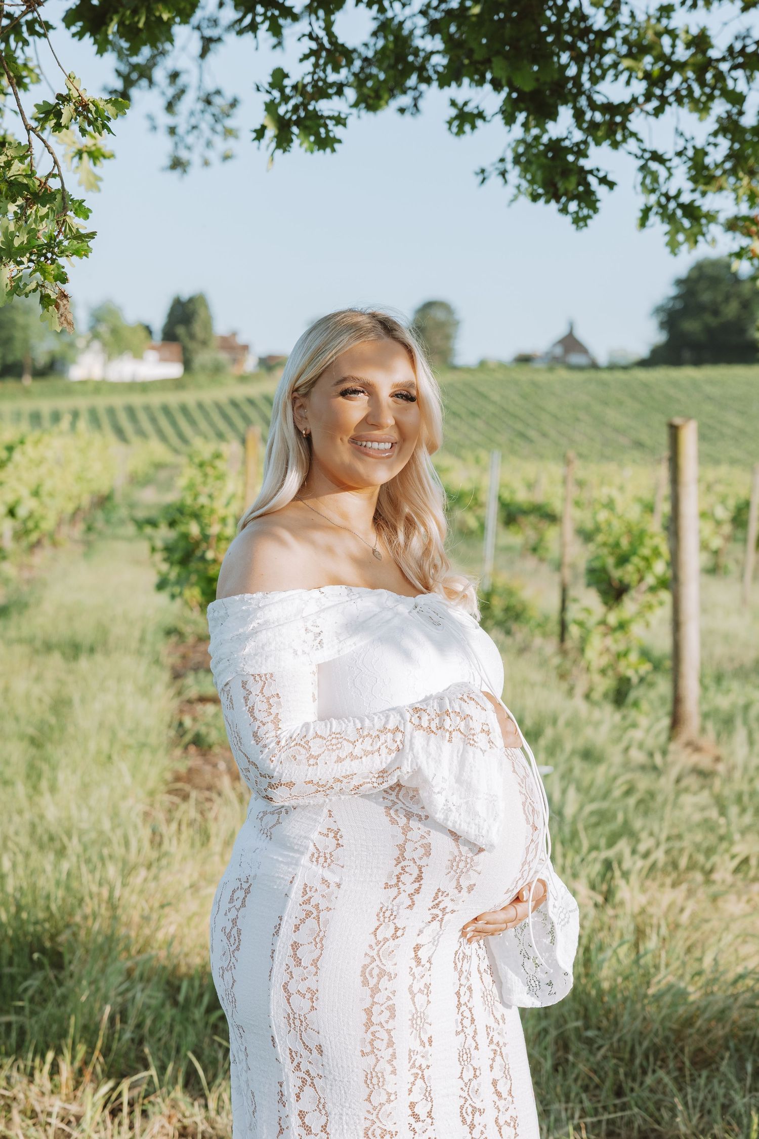 A pregnant woman in a white lace dress stands in front of a scenic vineyard on a sunny summer day.