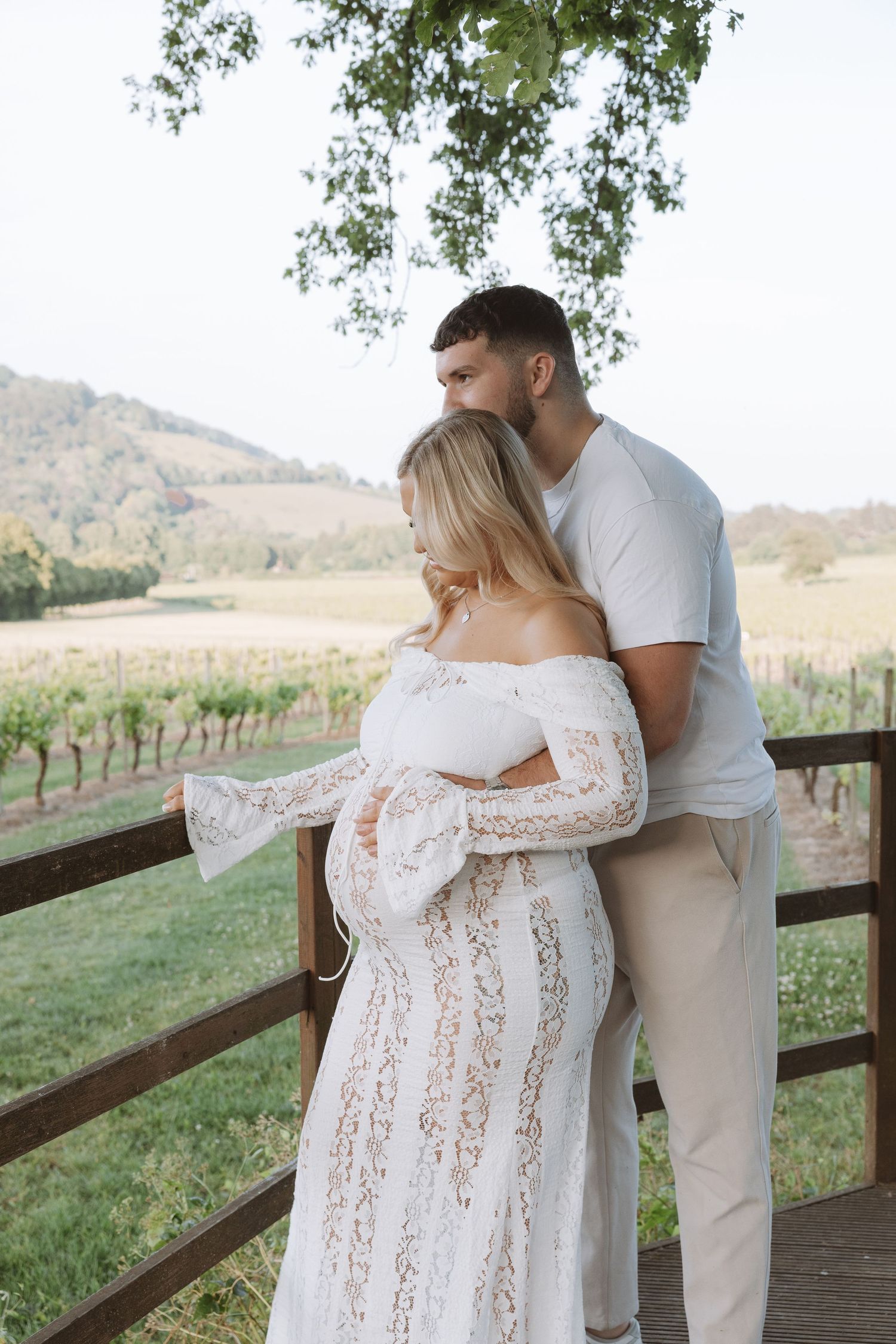 Expectant couple embraces on wooden deck overlooking vineyard and rolling hills in romantic maternity photoshoot.
