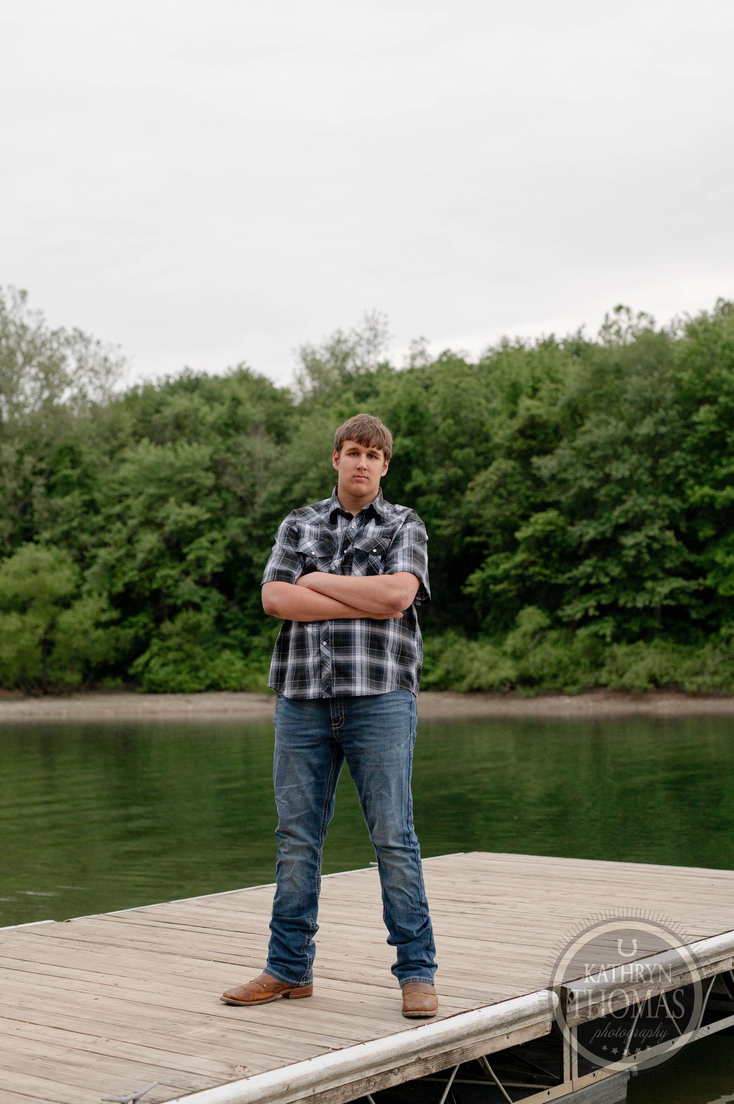A person in a plaid shirt and jeans stands confidently on a wooden dock overlooking a peaceful lake surrounded by trees.