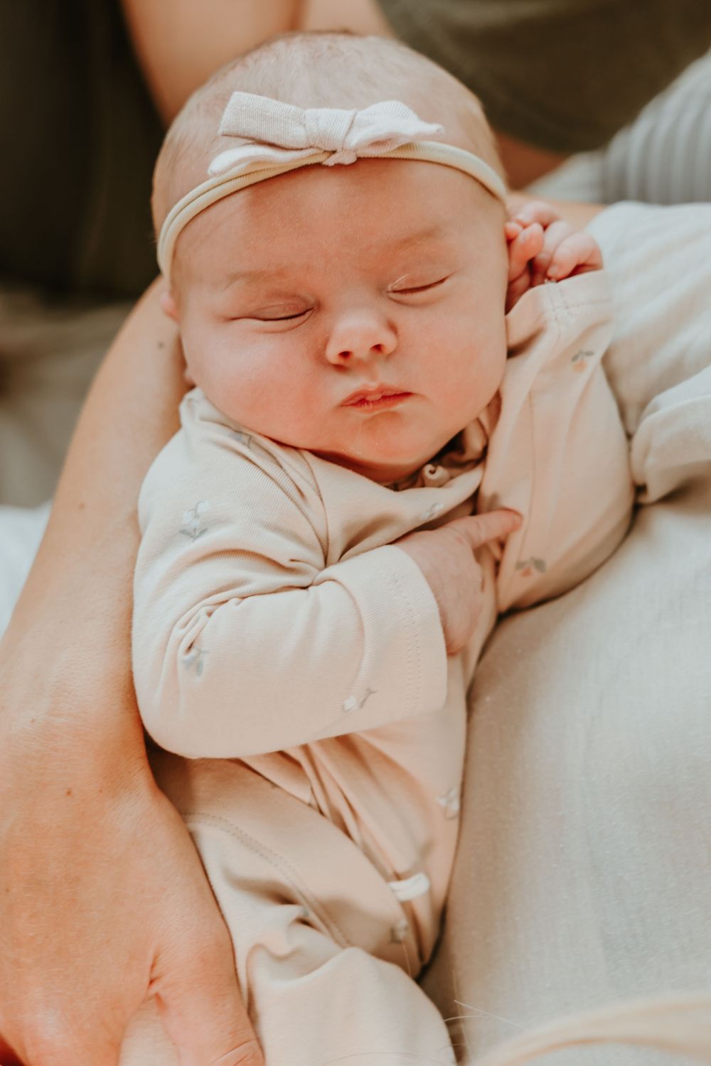 A sleeping newborn baby wearing a cream colored onesie and bow headband rests peacefully in someone's arms.