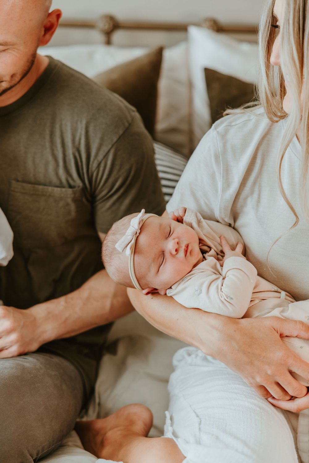A sleeping newborn baby wearing a white outfit and headband rests peacefully in their parents' arms on a couch.