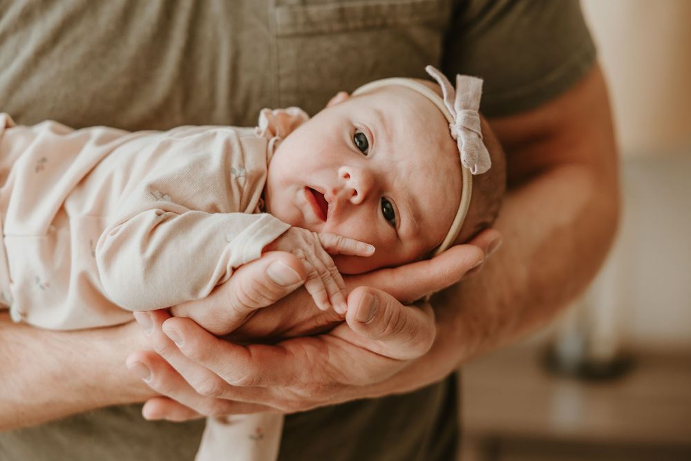 A small baby with a white bow headband rests gently in their parent's supportive hands.