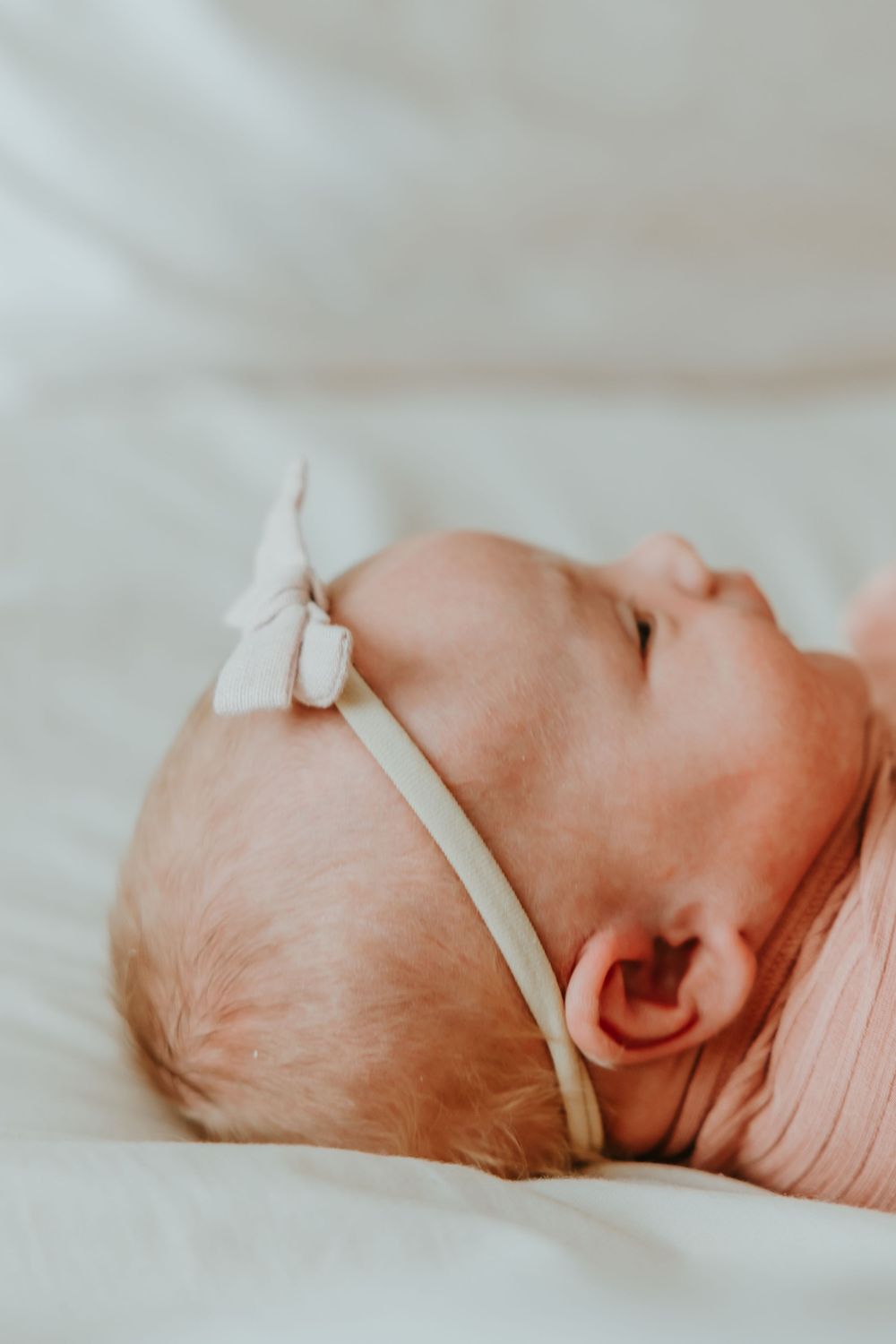 Close up of sleeping newborn baby in pink wrap wearing a delicate white bow headband against soft light background.