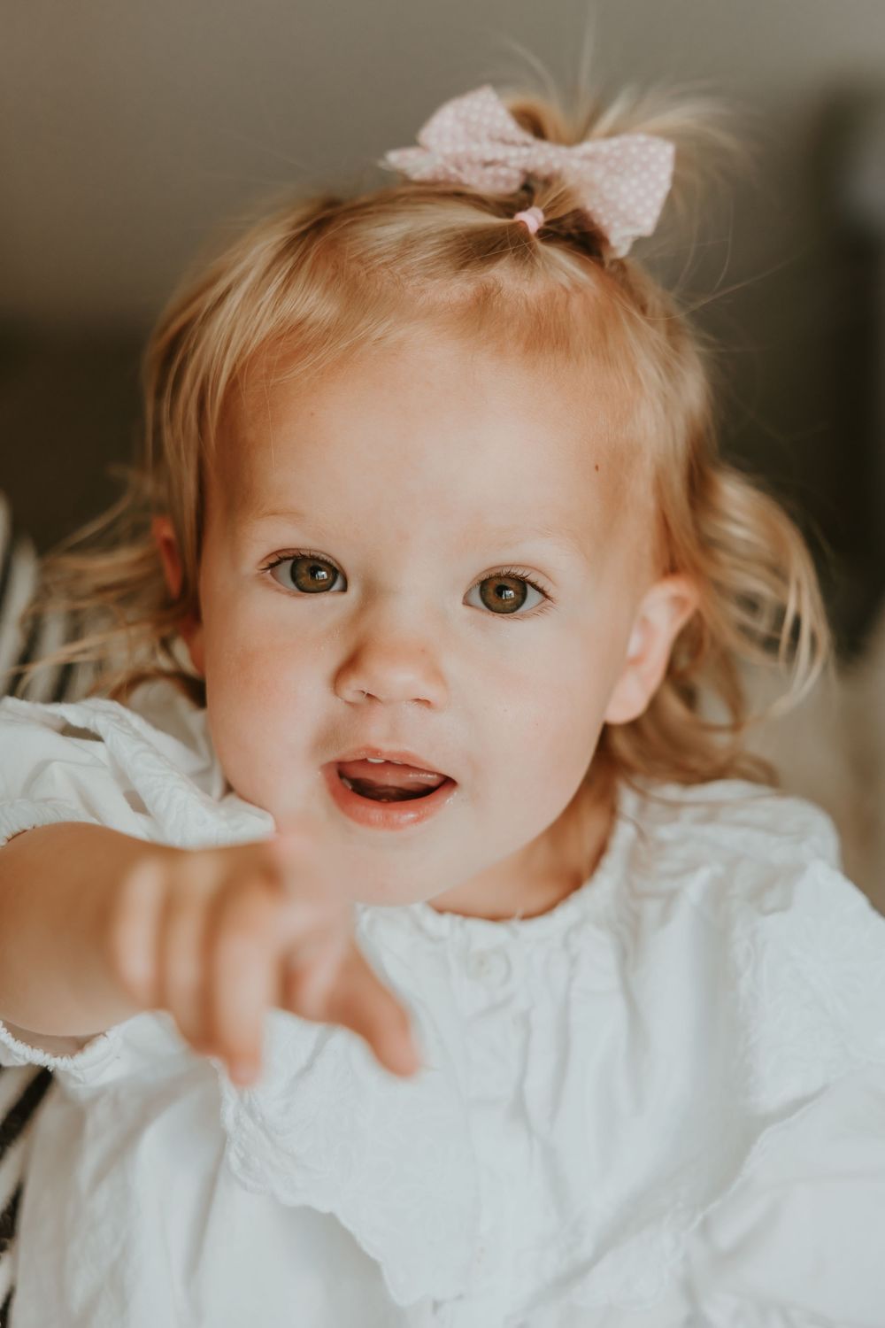 Young toddler with blonde hair wearing white dress and bow points towards camera with curious expression.